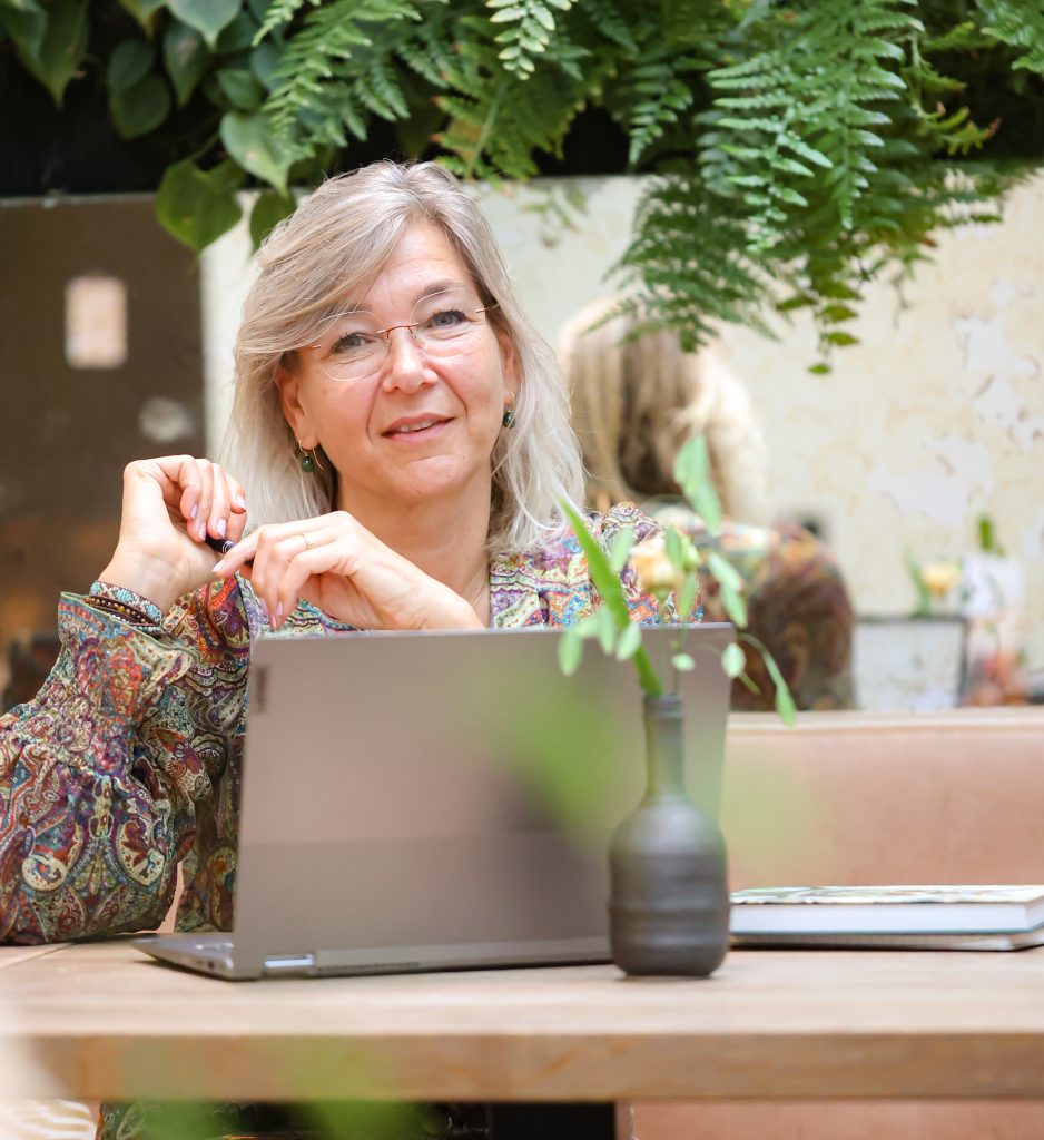 Melanie van De MEL Methode zit aan tafel achter haar laptop in een rustige, groene omgeving.